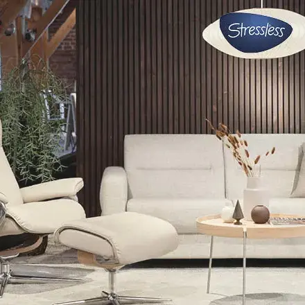 Heller Relaxsessel mit Hocker neben Sofa und Tisch, Logo Stressless oben rechts